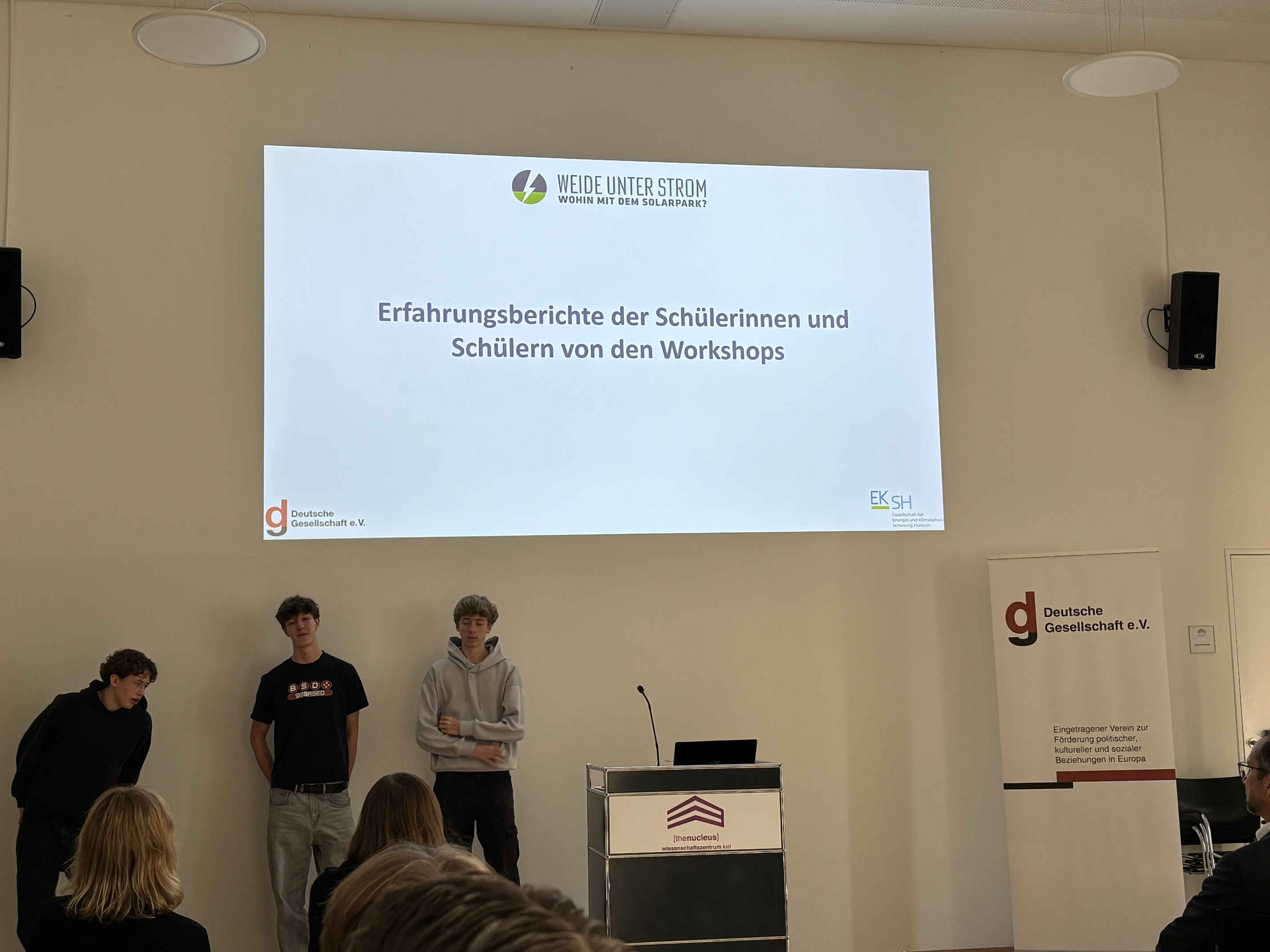 Drei Schüler stehen neben einem Pult und stellen ihre Ergebnisse vor.