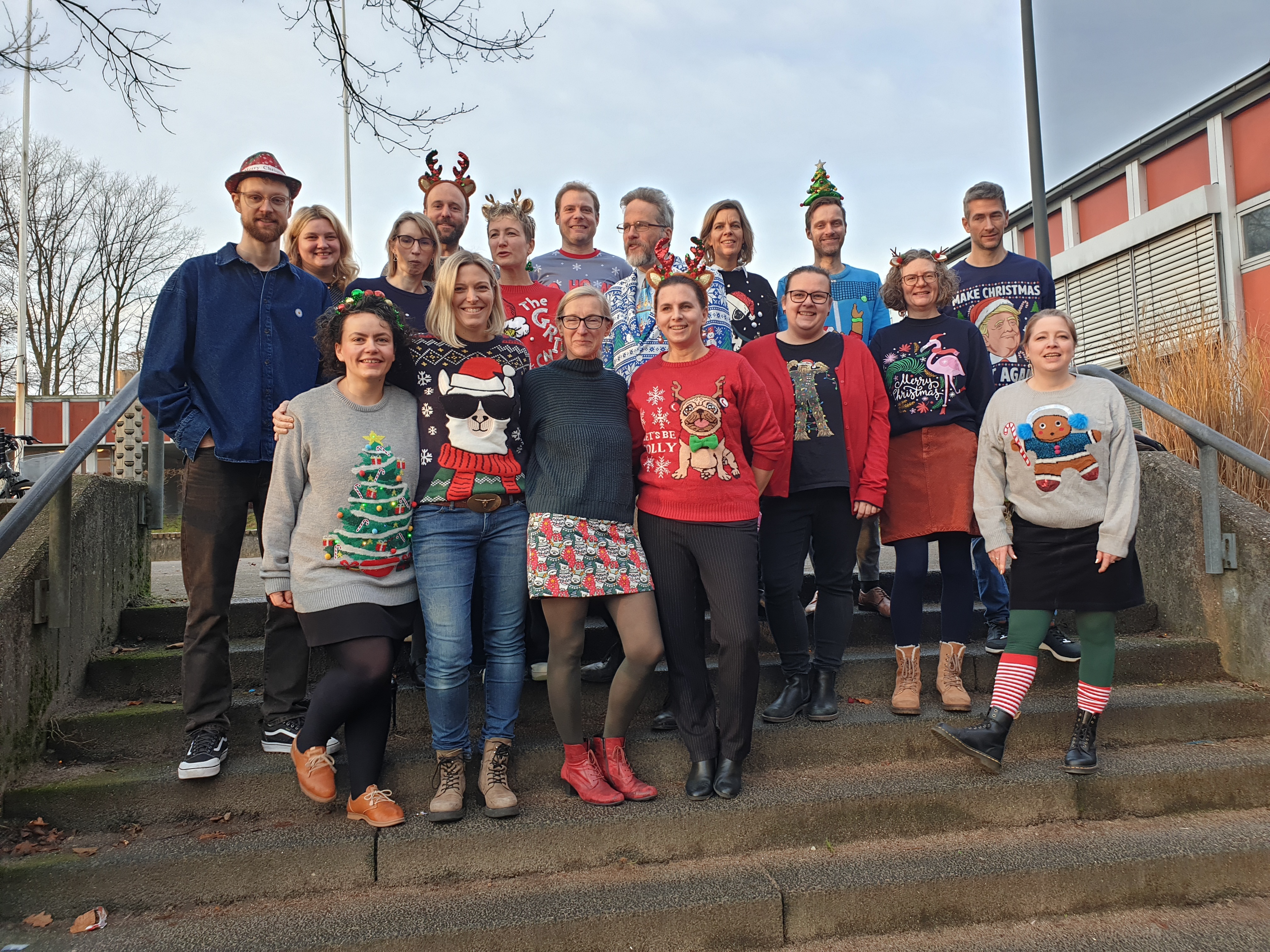 Ein Teil des Kollegiums steht auf der Treppe im Freien. Alle haben ugly christmas pullover an. Manche haben Rentierohren oder einen Weihnachtsbaum auf dem Kopf.