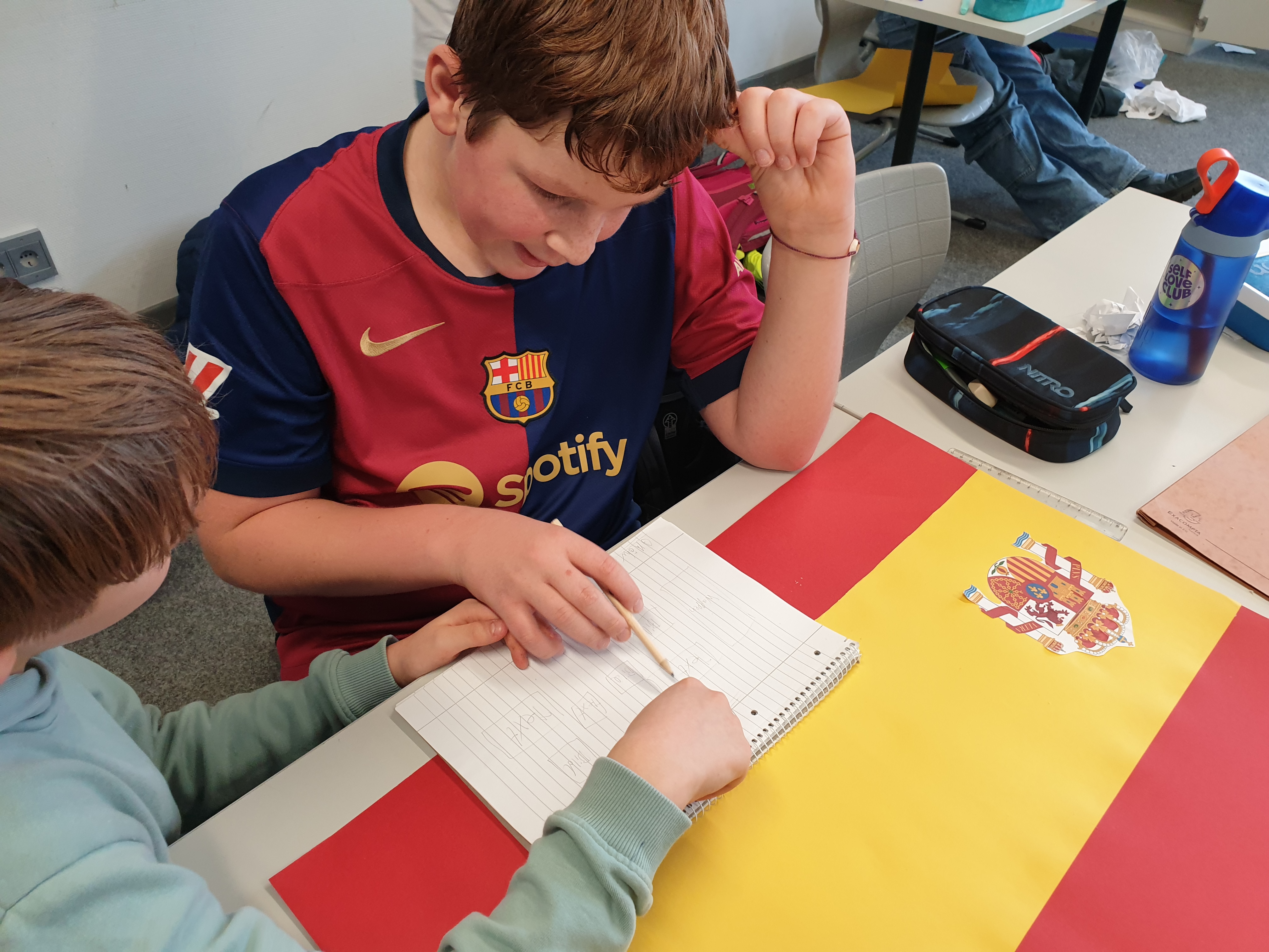 Zwei Jungen sitzen vor einem Plakat, dass wie die Flagge Spaniens aussieht. Sie diskutieren über die Plakatgestaltung.